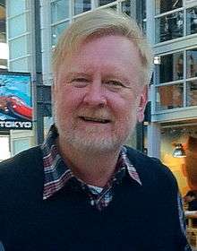 An older man is seen wearing a blue sweatshirt over a collared shirt with a red and blue plaid design.