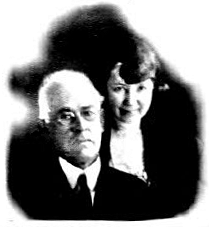 Image: A middle-aged Anglo couple, shown from the chest up: William, seated, is white-haired, wearing glasses and a dark suit and tie, and looks serious; Clara, standing behind him, is dark-haired and pleasant-faced, wearing what appears to be a dark cardigan over a white blouse.