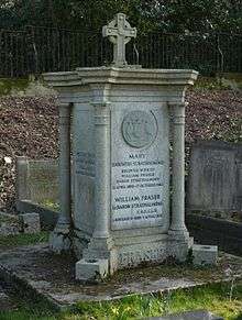 A granite, squared plinth with a column at each corner and a small cross on top of it, among other headstones