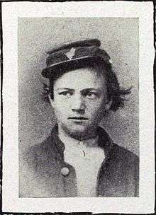 Head and shoulders of a young white man with unkempt hair sticking out from under a tilted forage cap. He is wearing an open jacket over a white shirt.
