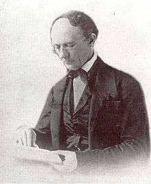 An older man with a receding hairline and reading glasses looks down at the book he is reading. He wears a dark suit with an old fashioned bow-tie