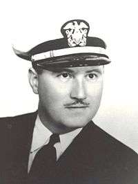 Head of a white man with a very thin mustache, wearing a dark jacket over a white shirt and dark tie and a white peaked cap with an eagle-shield-anchor emblem on the front.