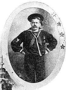 White man with mustache, hands on his hips, wearing a full sailor suit with lanyard around the neck, and single medal on the left breast, and a flat cap. Around the portrait is an oval-shaped frame with four stars.