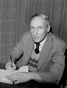 Older man wearing a chequered sweater under a suit coat. He is sitting at a table and signing his autograph in a book, facing the camera.