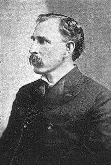 A man in his late forties with black hair and a mustache. He is wearing a tightly-buttoned black coat and facing left.