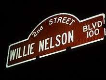 A sign of a street that reads "2nd street, Willie Nelson BLVD 100". It is night time and the sign is lighted. The borders and letters are white and the inside is red.