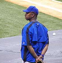An African-American man, wearing a baseball hat and jacket, stands outside a baseball diamond.