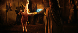 A little man in a hooded cloak with his back to the camera holds a lightening wand toward a two-legged animal that appears to be part goat and part ostrich.