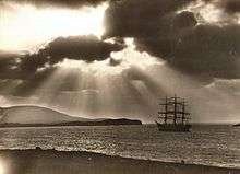 A sepia-tone photograph of a 3-masted 19th century sailing vessel at sea with the shore in the foreground and dark hills beyond. The sun's rays cast light and shadows in the sky as they break through the clouds.