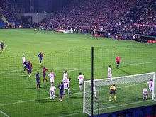 A photograph from behind the goal shows Barcelona and Wisła Kraków players gathering in the near penalty area for a corner kick. Wisła Kraków players are standing closer to the goal, and the referee is standing on the edge of the penalty area.