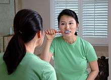 An Asian woman brushing her teeth while looking in a mirror.