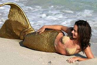 A woman wearing a golden costume mermaid tail and a bikini lies on the beach.