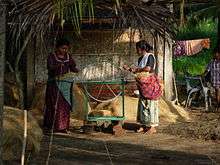 Two women spinning coconut fibre on small green loom outside a home