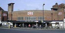 A brown-bricked building with a rectangular, dark blue sign reading "WOOD GREEN STATION" in white letters all under a light blue sky