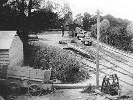 A single railway track curves sharply past a short platform with a small wooden hut behind it. A lone person stands on the platform. In front of the hut is a small shed with a very large overhanging roof, with another railway track terminating under the roof. An overgrown railway track diverges from the other two tracks, terminating at a set of buffers near another wooden hut surrounded by farming machinery and an apparently derelict horsecart. The tracks and buildings are all sited in a small triangular clearing, which is completely surrounded by trees.