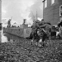 British soldiers assist a wounded soldier between buildings towards the sea and a waiting landing craft left