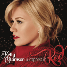 A blonde haired woman whose neck is wrapped in a red scarf looking sideways against a dark background; below her, the word-marks "Kelly Clarkson" and "Wrapped in" are printed in stylized "Feel Script" and "Bree" typefaces aside a red ribbon-styled "Red" word-mark.