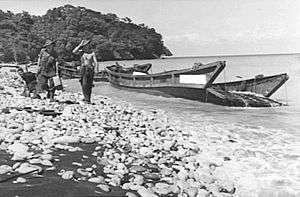 Two soldiers walk along a beach beside the wrecked remains of a number of barges