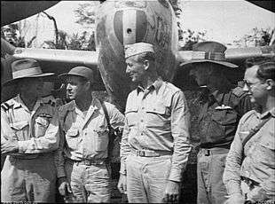 Five men in uniform stand in front of a dual propeller aircraft.