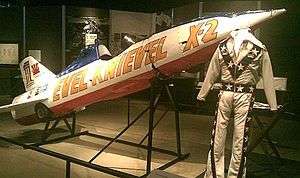 Rocket with front end tilted upwards and a flight suit in front of it