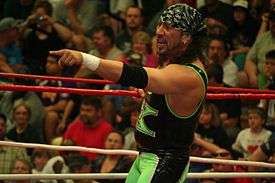 An adult male with long black hair wearing green and black wrestling gear standing in a ring pointing away from himself.