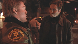 A man in a sheriff's uniform is talking to a man with black hair dressed in brown jacket. The film quality is deliberately low-quality.