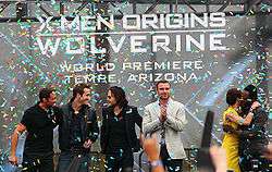 Atop a stage are three man in black clothing, Liev Schreiber wearing a gray jacket and black pants, and Lynn Collins, wearing a yellow dress, hugging will.i.am, who is in black clothing. In the background is a billboard reading "X-Men Origins Wolverine: World Premiere – Tempe, Arizona. Colored paper flies through the stage.