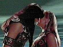 On the left is a tall black haired woman, dressed in leather. She is bending down to kiss a young, slightly shorter, blonde haired woman.