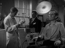 A black-and-white screenshot from the film depicting a tall man in a white coat reading a clipboard watched by a man with a moustache wearing a dark suit. In the foreground a man is seated rigidly staring straight ahead, his eyes and face sunken into a skeletal appearance