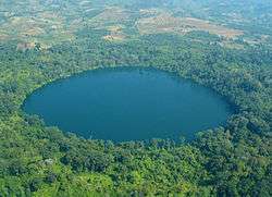 A deep blue, round lake surrounded with forest. Nearby, the forest has been replaced with fields.