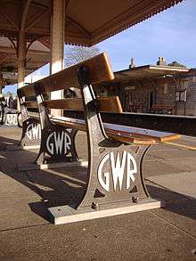 A bench seen from low down and behind. Three brown-painted cast iron legs have "G W R" cast into them in a circular mofif and painted white, and support two pairs of widely spaced wooden planks that form the seat and back.