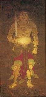 Frontal portrait of a frightening deity dressed only in a skirt-like garment holding a sword in his right hand.
