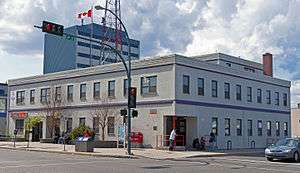 A two-story light gray building seen from across an intersection over which a traffic signal is displaying a red light. A Canadian flag is flying from the flat roof. There are taller buildings behind it.
