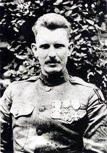 Head and shoulders of a man in military uniform with combed back hair and a neatly trimmed mustache. Three medals hang from ribbons on his chest.