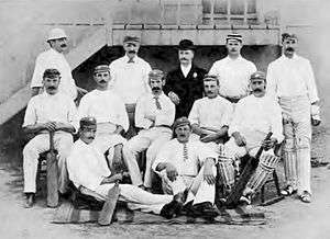 A cricket team arranged in three rows. Apart from one man in a suit, they are all wearing cricket whites.