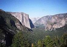 A glacial-carved valley filled with evergreen trees has a hanging valley and waterfall on the right and high cliff face on the left.