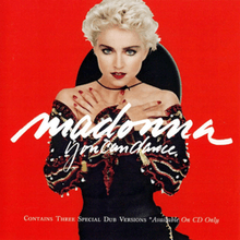 Madonna with short, blond hair, wearing a black dress, with her hands folded against her breasts and standing in front of a red background. She has a Spanish hat attached to her neck.