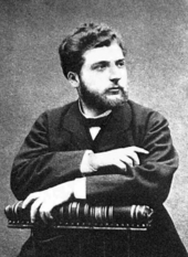  A young man, lightly bearded, sits in a posed position, facing half-right. He is formally dressed in dark clothes, and is holding a large book or scroll