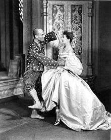 A barefoot man in Asian-style dress dances exuberantly with a woman in a formal gown with a large hoop skirt