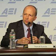 A man with close-cropped, receding hair,wearing a suite, looking intently slightly to his right. He is sitting at a table with a microphone against a blue, repeating ARI logo.