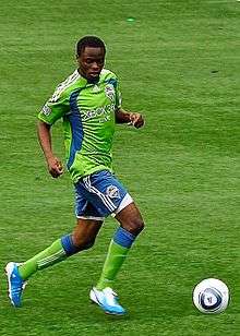Zakuani dribbles a ball down the field while wearing a Sounders kit
