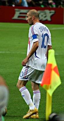 A bald-headed man wearing a white shirt and white shorts with the number "10" displayed on the back of the shirt.