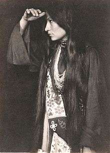 Photo of Zitkála Šá in profile, wearing Native American dress, with long dark hair hanging below waist, holding hand at forehead and looking into the distance