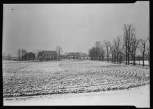 A black-and-white photograph of a cornfield in the foreground and farm buildings in the background during Winter.