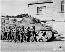 A tank is parked in front of a house. Seven soldiers stand in front of the tank.
