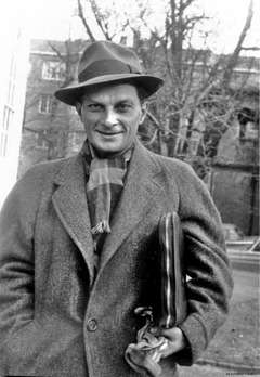 A smiling man in a hat and heavy winter coat and scarf, carrying a portfolio tucked under his arm