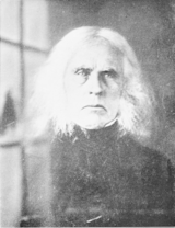 An elderly man with black clothing and white hair down to his shoulders.