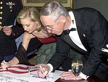 A young woman and an older man, both dressed in formal wear, leaning over to sign a poster.